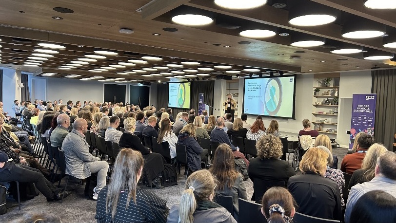 A full conference room at the Global Payroll Alliance Summit 2025, delegates seated facing the stage as Dannielle Haig presents beside her title slide
